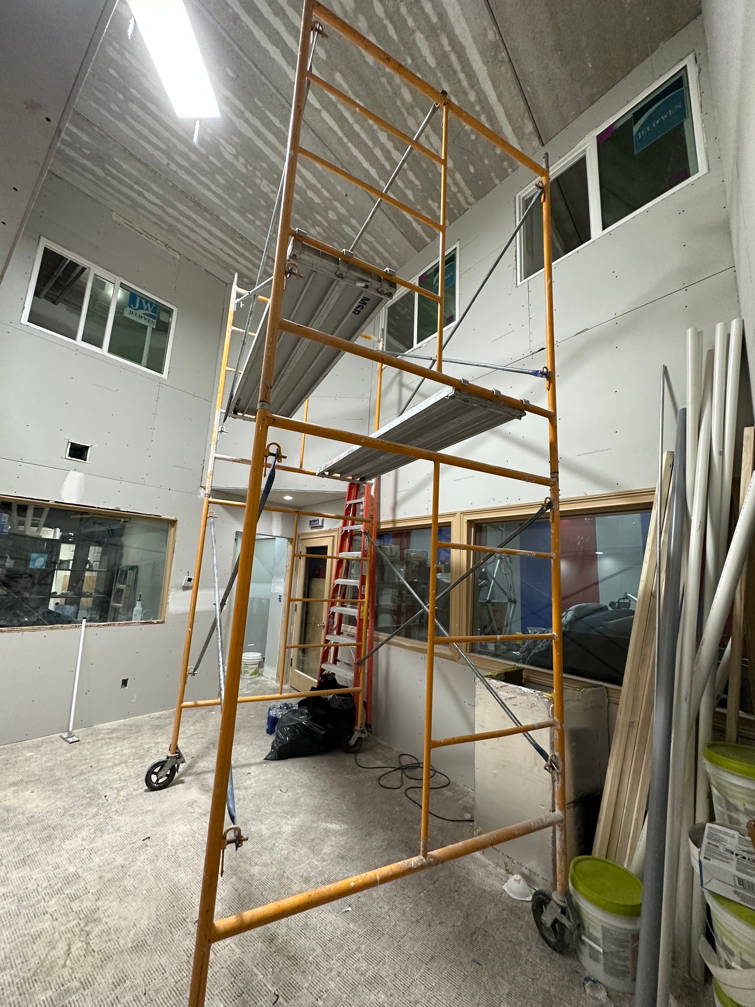Yellow metal scaffolding inside a room with unfinished drywall and installed windows under construction.
