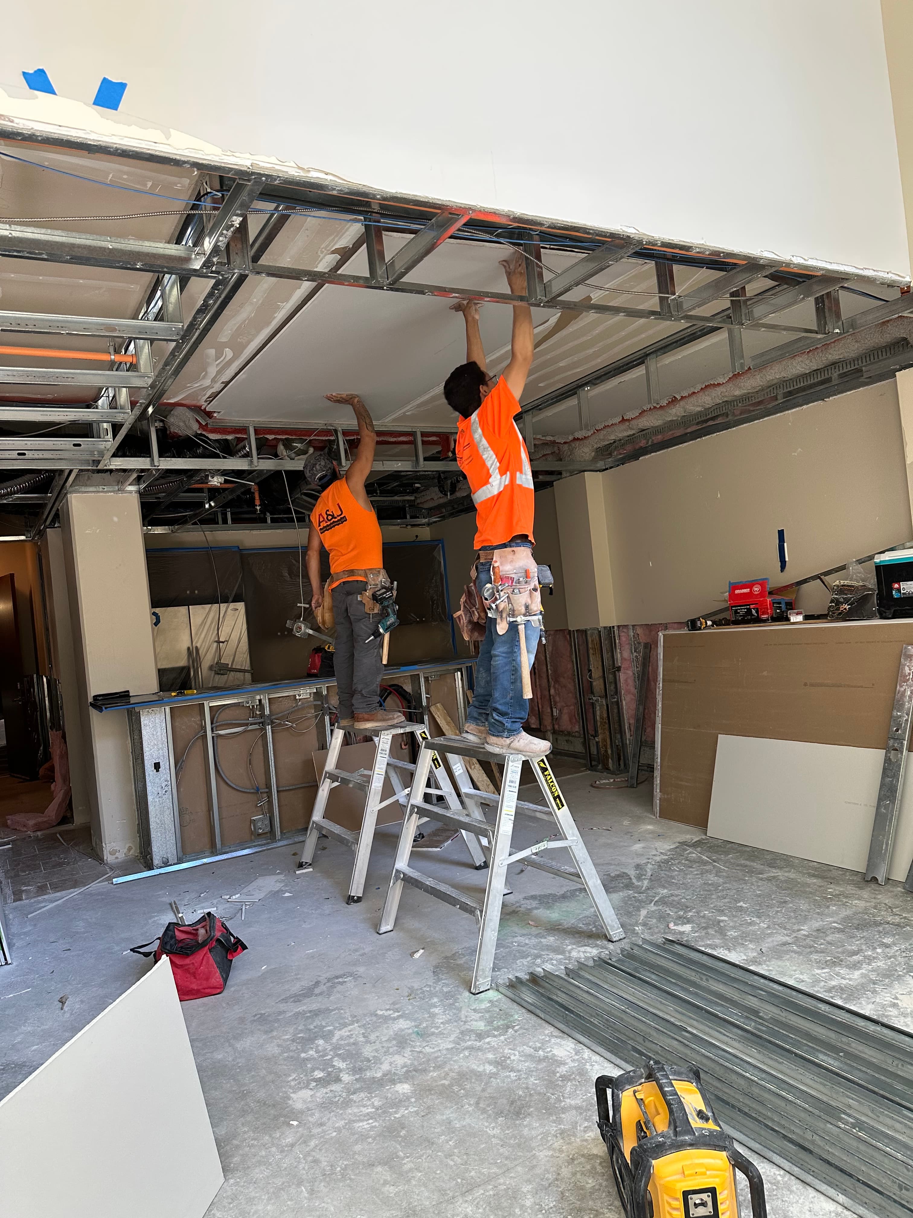 Two construction workers in orange shirts install drywall panels onto a metal ceiling frame.